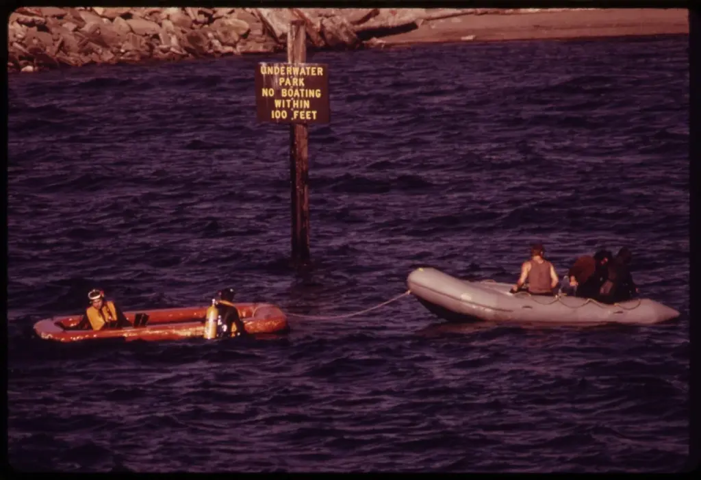Divers in drysuits in small inflatable boats beside a sign at Edmonds Underwater Park, Washington
