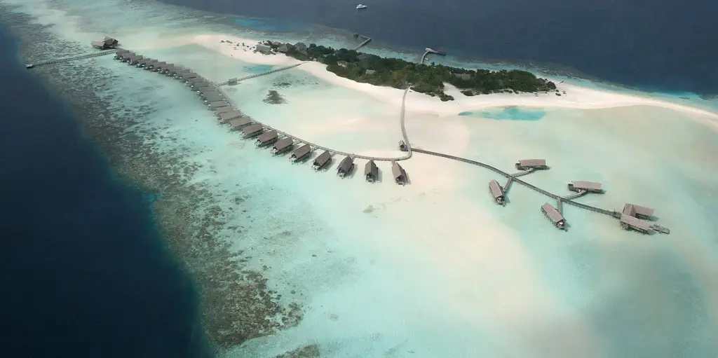 Aerial view of a small Maldives resort island with a long line of overwater villas over a white-sand reef flat